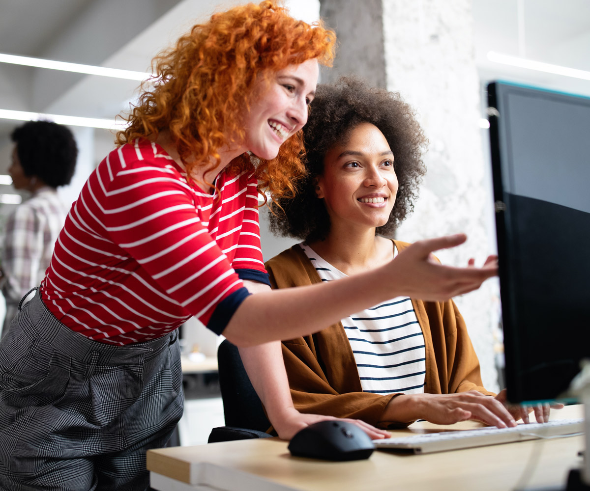 Two women are sitting at a desk and one is using a computer.